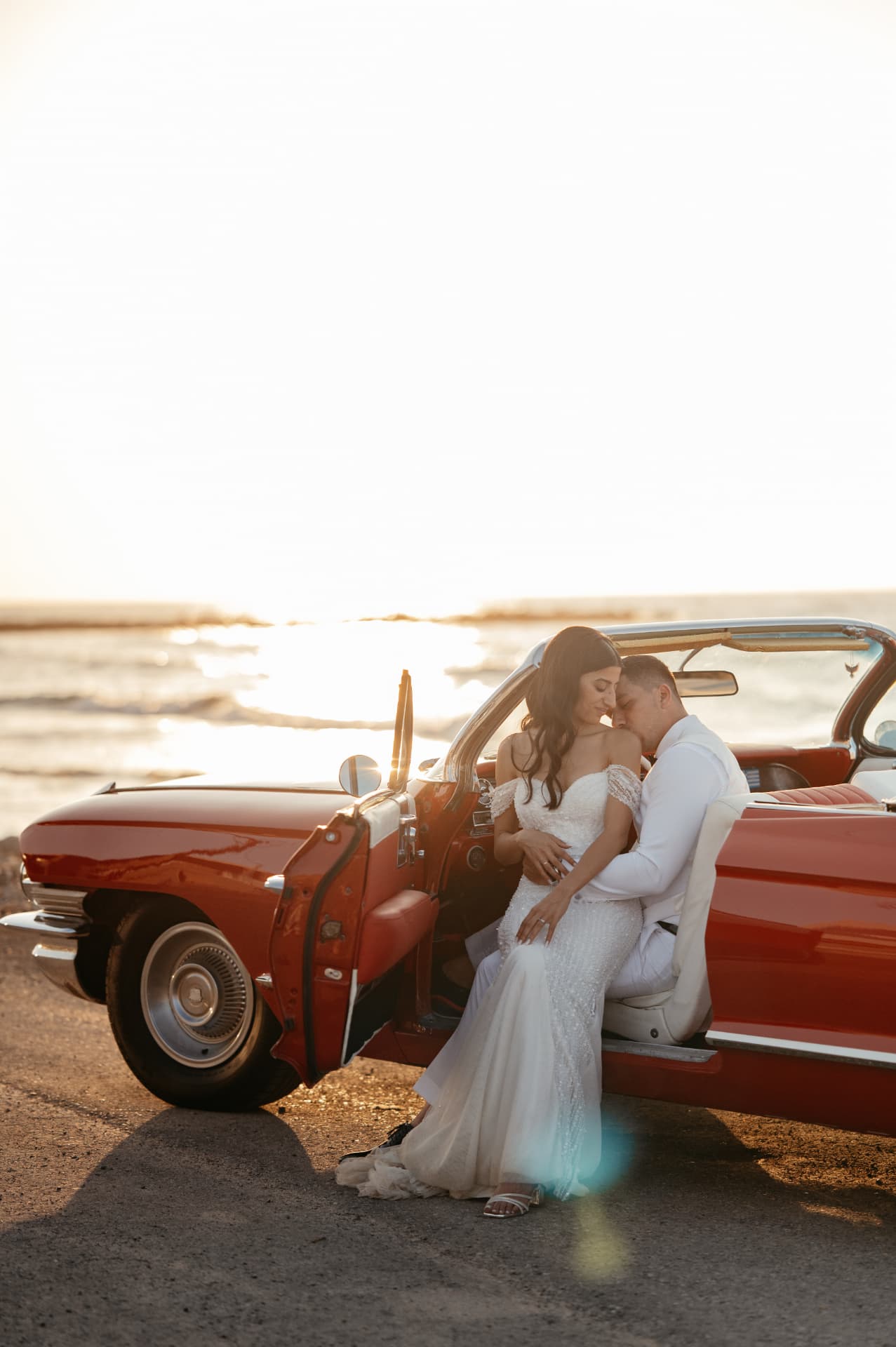 A groom kissing his bride's shoulder in a red vintage convertible