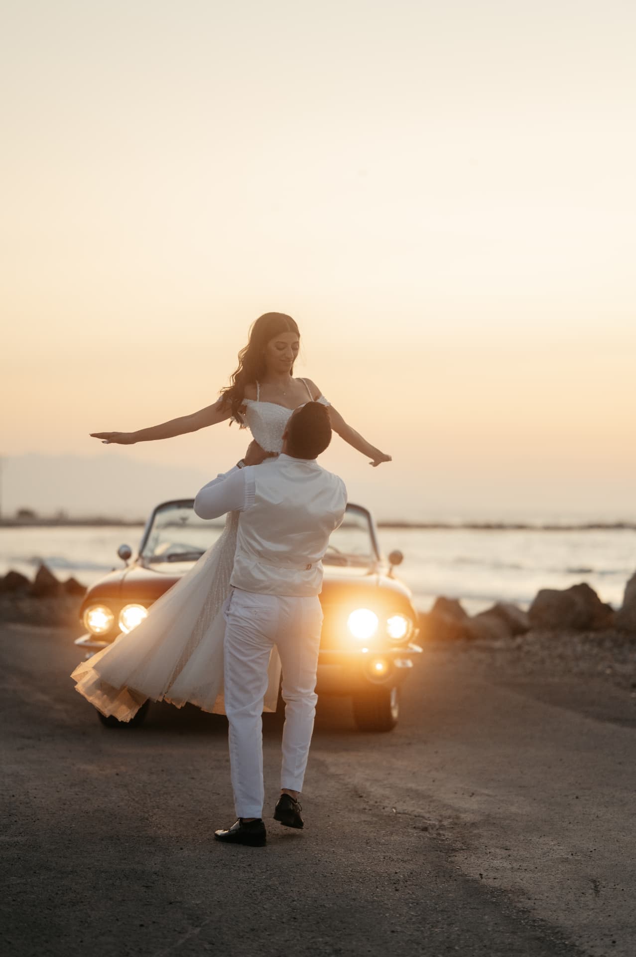 A groom lifting his bride in front of a vintage car at sunset